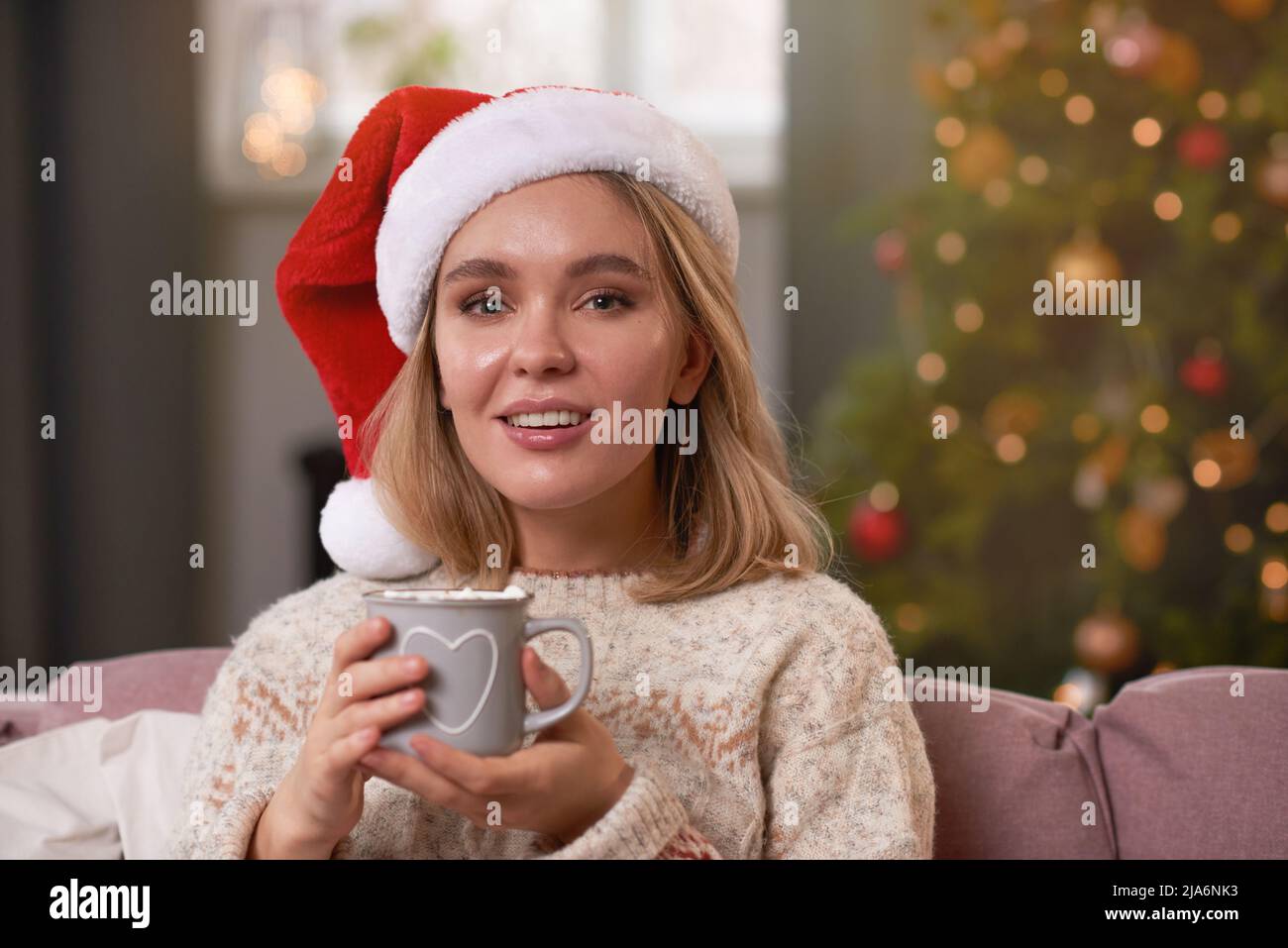 Mittellange Nahaufnahme einer attraktiven jungen Frau mit Weihnachtsmann-Hut, die eine Tasse mit heißem Kakao und Marschmalow in der Hand hält Stockfoto