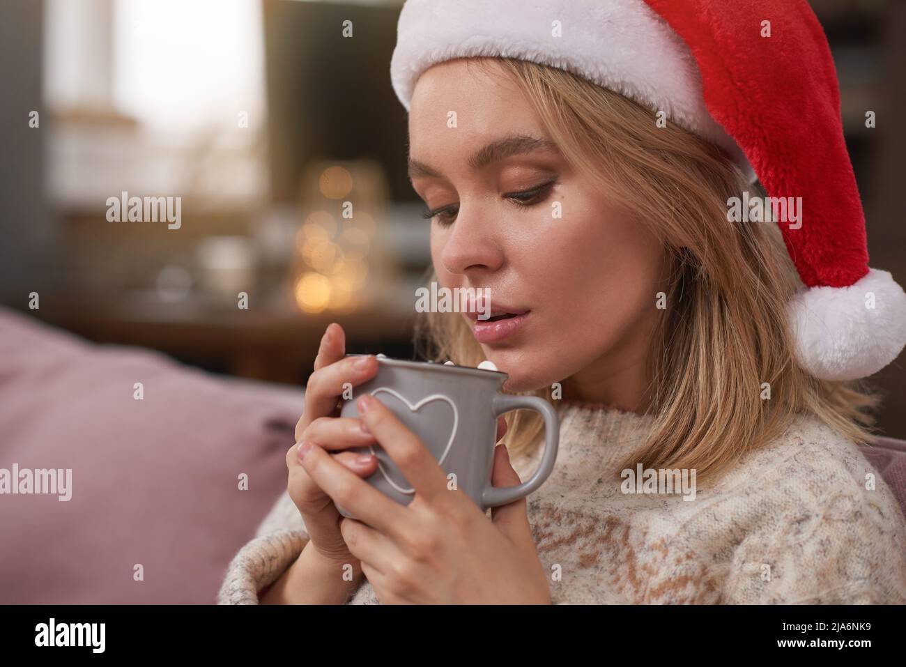 Nahaufnahme einer blonden kaukasischen Frau in warmem Pullover und Weihnachtsmütze, die am Weihnachtsabend heißen Kakao mit süßem Marschmalow trinkt Stockfoto