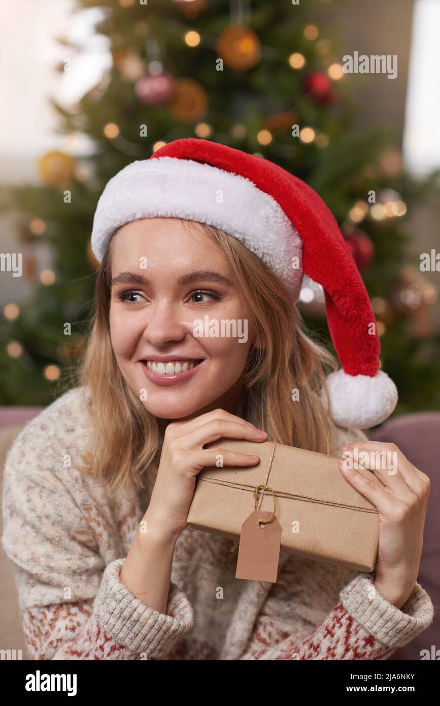 Mittellange Nahaufnahme Porträt einer glücklichen jungen Frau in warmen Kleidern und Weihnachtsmütze mit Weihnachtsgeschenk, die wegschaut Stockfoto