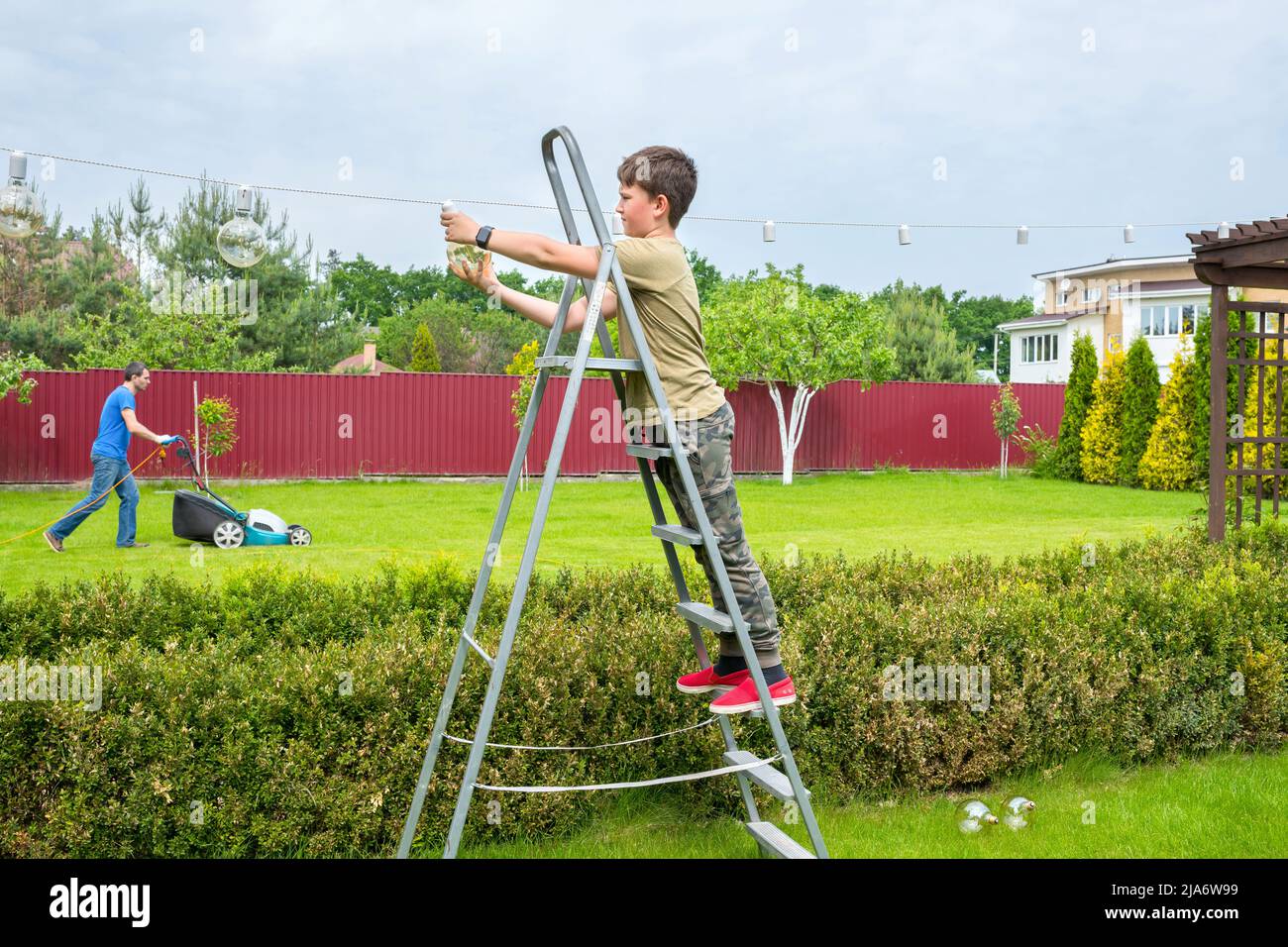 Ein Junge auf der Leiter steckt Glühlampen im Hinterhof an. Vorbereitung auf den Urlaub. Eine Familie putzt den Garten. Stockfoto