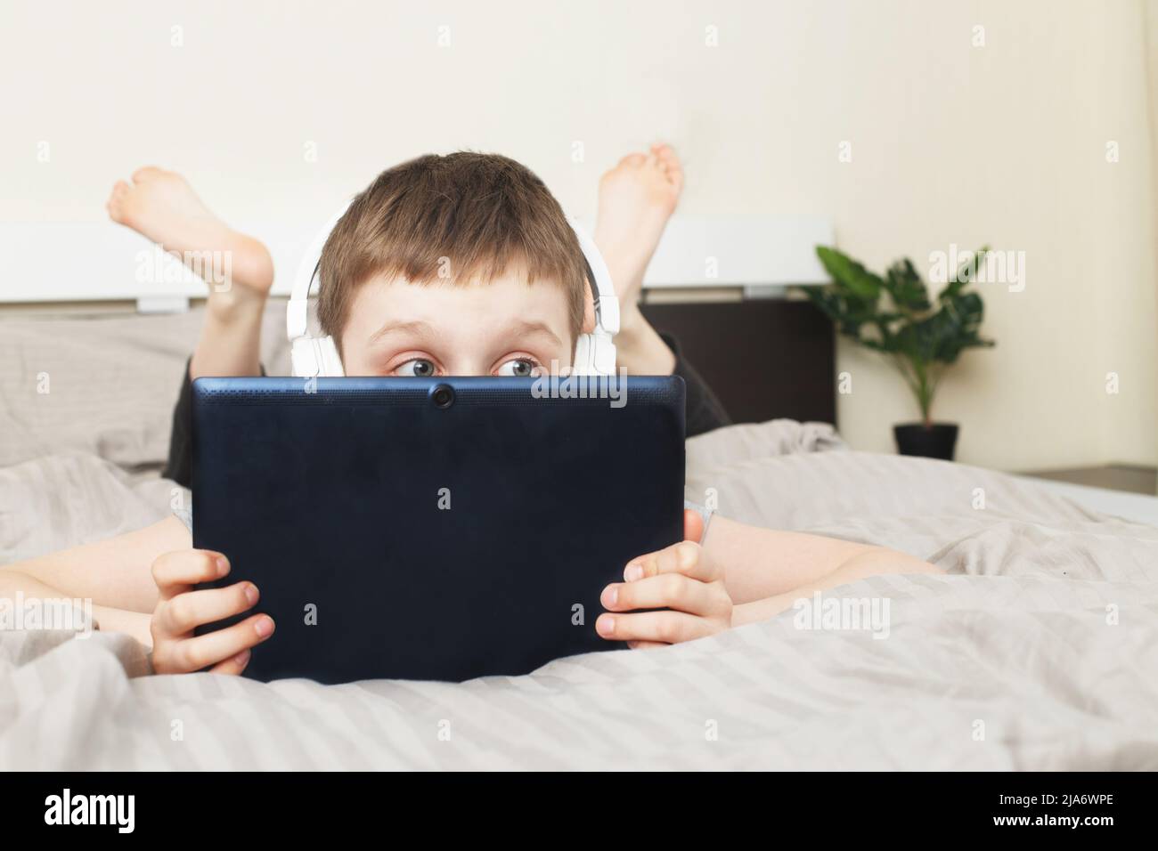 Der Junge, der zu Hause auf dem Bett lag, bedeckte sein Gesicht mit einem pc-Tablet und las online, lernte oder hörte Musik mit kabellosen Kopfhörern Stockfoto