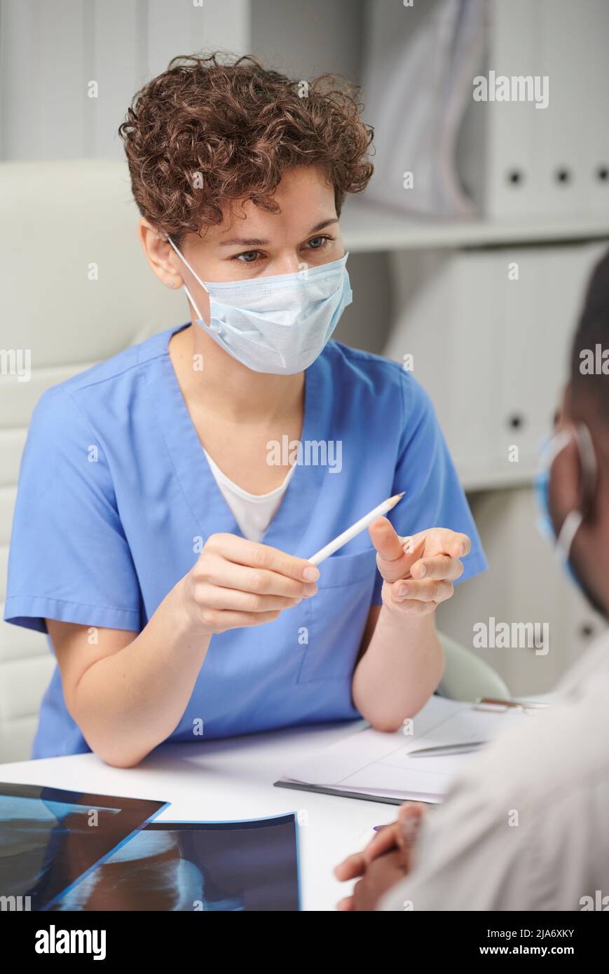Vertikale Aufnahme über der Schulter einer nicht erkennbaren Frau mit kurzen Haaren, die eine Schutzmaske auf dem Gesicht trägt und den Patienten konsultiert Stockfoto