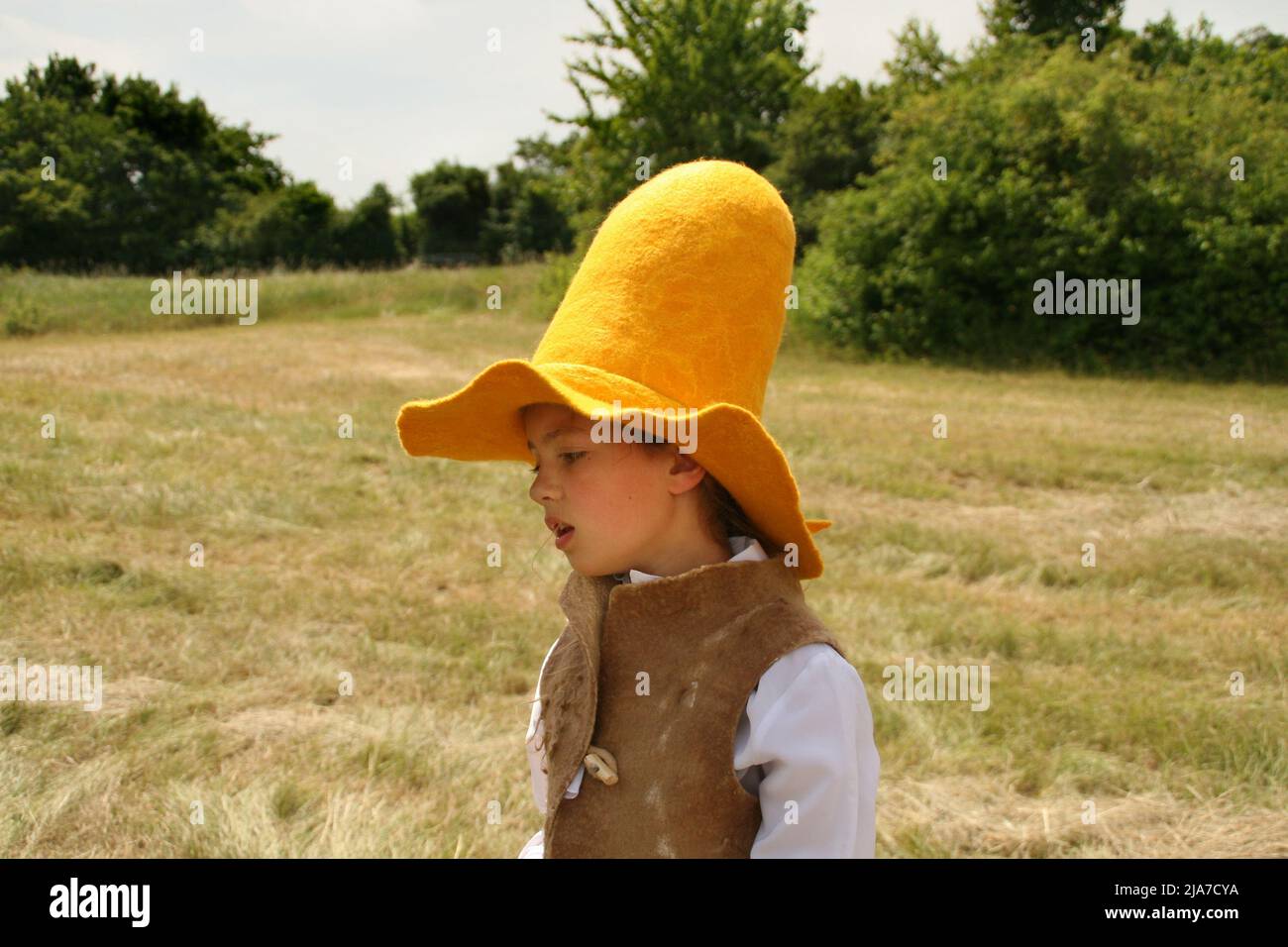 Mädchen in einem hohen gelben Bauernhut mit dem breiten Rand läuft im Sommer auf dem grünen Feld. Sie hat ein langes braunes Haar, ein einziges Zopf hängt herunter Stockfoto