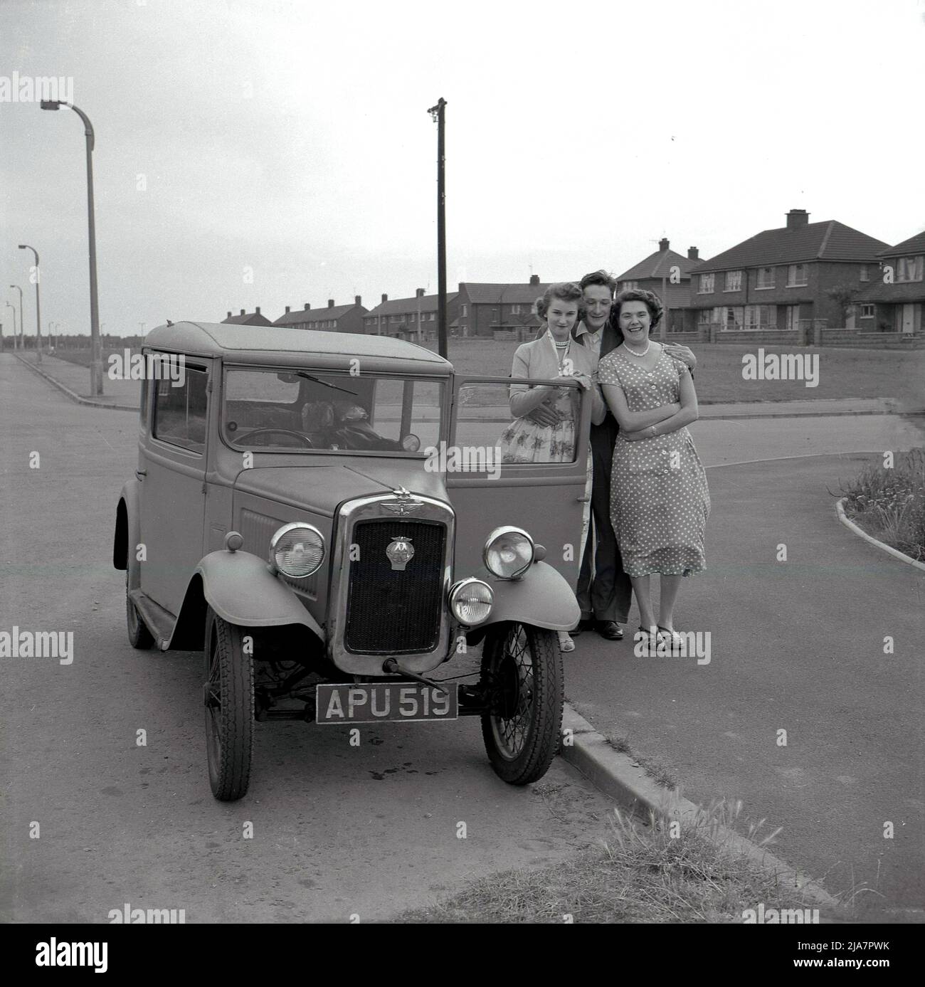 1950s, im historischen Großbritannien der Nachkriegszeit und auf einem Bürgersteig in einer Wohnsiedlung, stehen ein junger Mann, seine Freundin und Mutter zusammen für ein Foto von ihrem Auto, einer Austin Seven. Der britische Made Austin 7 war ein kleines - mit dem Spitznamen "Baby Austin" - Economy-Auto, das von 1923 bis 1939 produziert wurde und das Fahrzeugeigentum an diejenigen brachte, die es sich nie zuvor leisten konnten, und beworben als: "Ein Penny-a-Mile mit vier mehr deckt die laufenden Kosten". Es wurde auch Frauen als „das Auto für die weibliche Note“ beworben. Der Wagen war in Großbritannien bis in den 1960s noch im Einsatz. Stockfoto