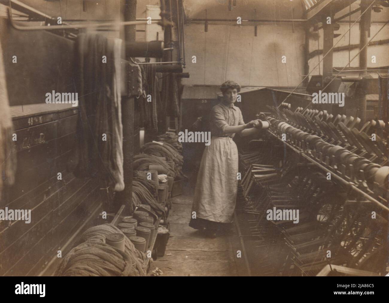 Viktorianisches Foto einer Frau in einer Schürze, die neben Maschinen in einer Wollmühle von Bradford steht, sind Wollkeins an der Wand hinter dem Mühlenarbeiter aufgereiht. Das Foto (eine Kabinettkarte) wurde von Joseph Foulds, 16 Church Street, Bradford, West Yorkshire, angefertigt Stockfoto