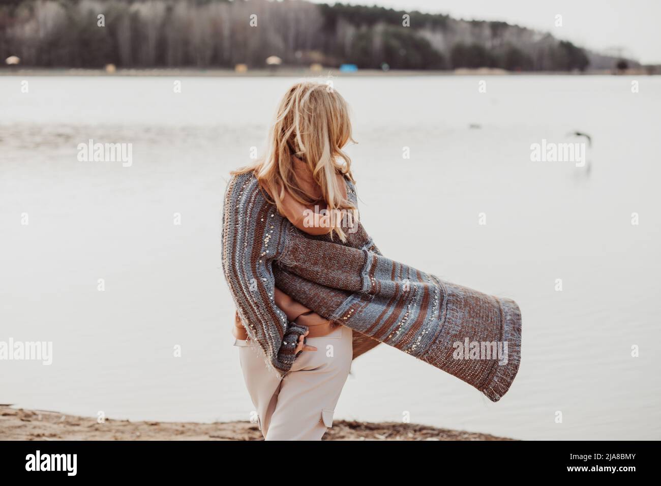 Porträt einer Frau mit langen, welligen, hellen Haaren, die eine braune Wolljacke und eine beige Hose trägt, die vom Sandstrand weg geht. Stockfoto Porträt einer Frau mit langen, welligen, hellen Haaren, die eine braune Wolljacke und eine beige Hose trägt, die vom Sandstrand weg geht. Stockfoto