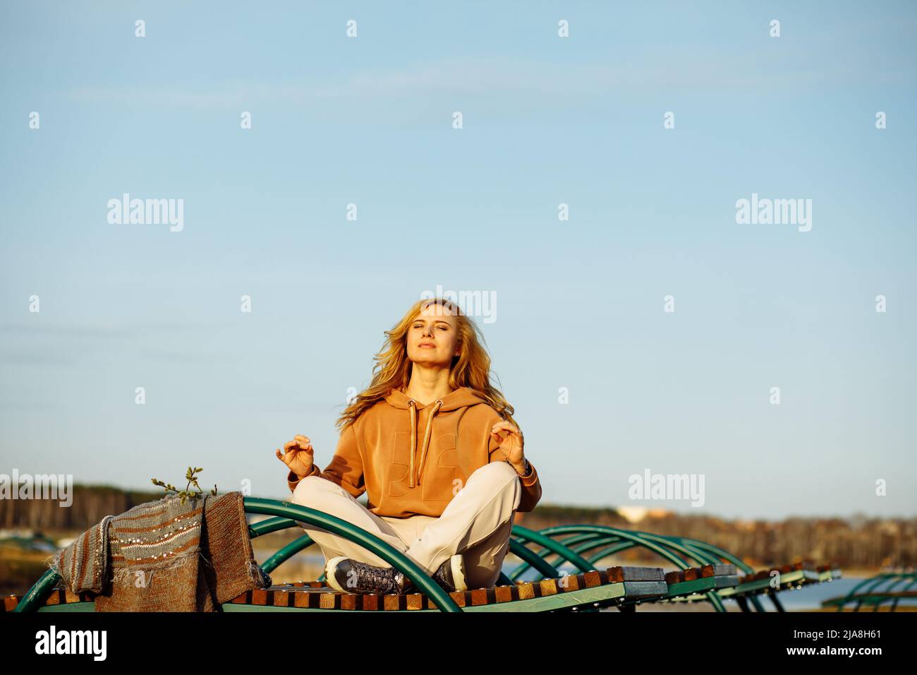 Porträt einer jungen Frau, die in Lotuspose mit Mudra-Geste auf der Lounge sitzt und das Gesicht durch geschlossene Augen gegen die Sonne ersetzt. Stockfoto