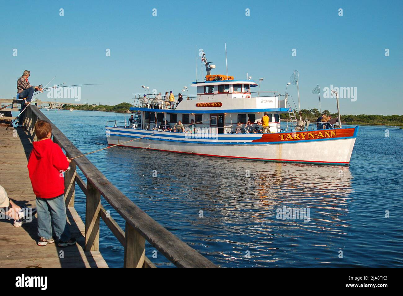Ein Charterboot fährt an einem kleinen Kind und seinem Vater vorbei und fischt von einem Pier im Captree State Park auf Long Island aus Stockfoto