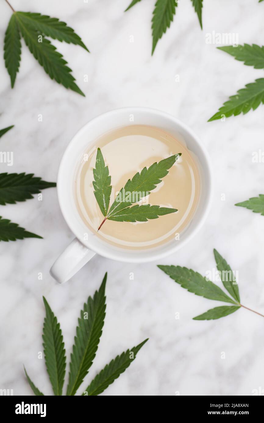 Weißer Becher mit Marihuana-Infusion mit grünem Blatt innen auf Marmortisch mit Blättern. Draufsicht. Stockfoto