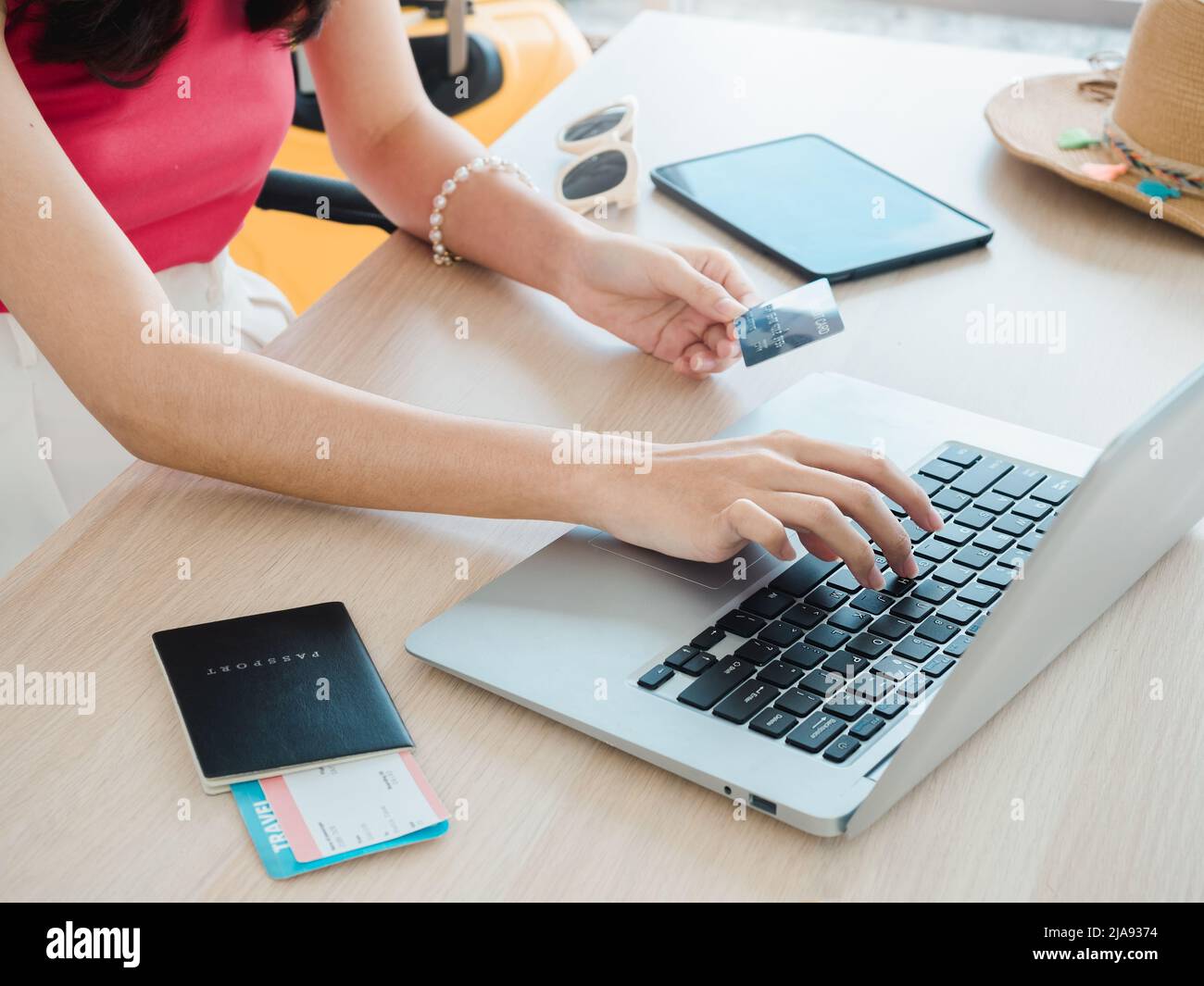 Die Hand der jungen Frau hält Kreditkarte und Reisepass, während sie den Laptop für die Flugbuchung benutzt, das Hotelzimmer für die Sommerreise auf dem Tisch, bereit für die Reise Stockfoto