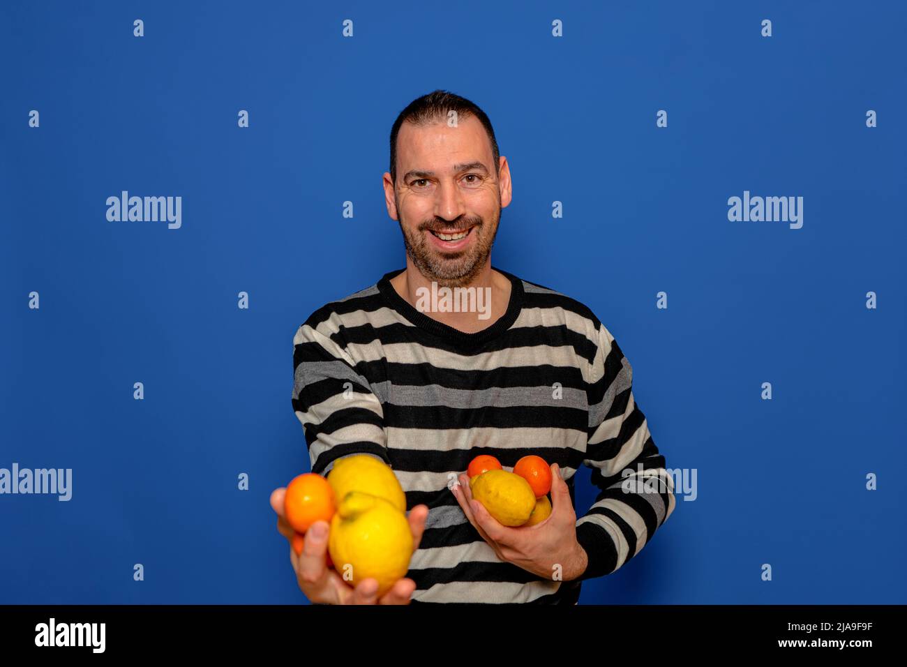 Bärtiger hispanischer Mann, der einen gestreiften Pullover trägt, bietet aufgeregt Orangen und Zitronen an, die er isoliert im blauen Studio in seinen Händen hält Stockfoto