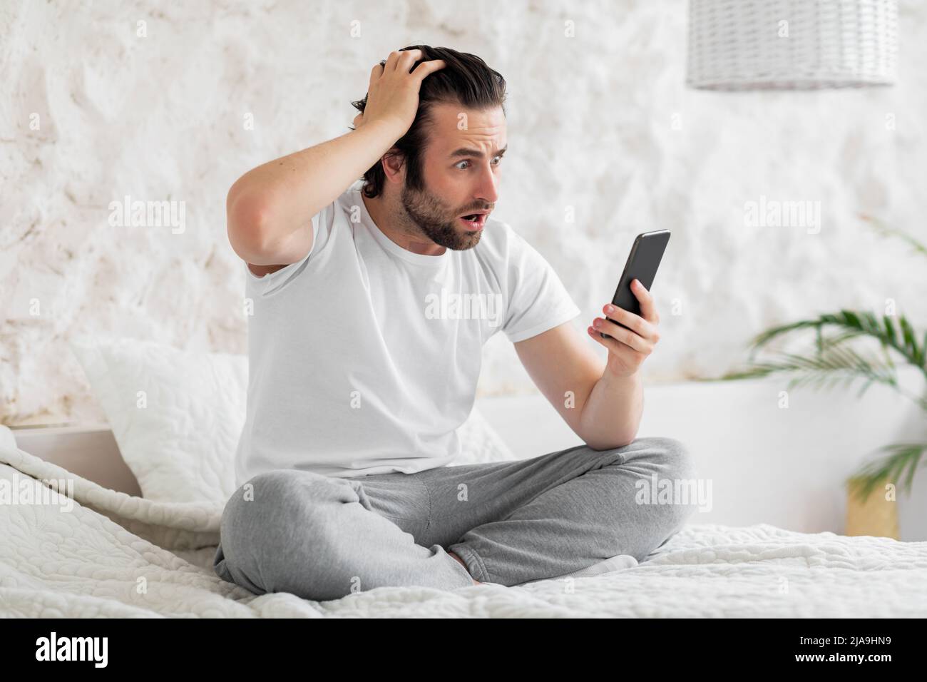 Schockierter junger Mann, der mit dem Smartphone auf dem Bett saß und den Kopf berührte Stockfoto