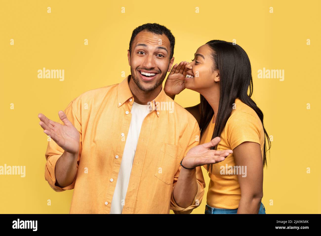 Junge schwarze Frau flüstert ihrem Freund ins Ohr und teilt Klatsch oder Nachrichten auf gelbem Studiohintergrund Stockfoto