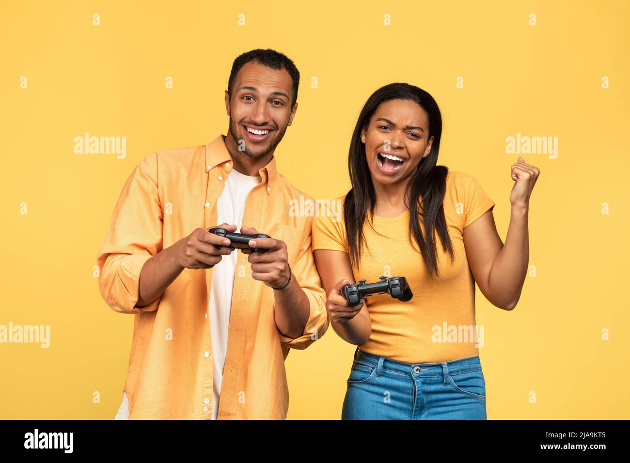 Aufgeregt junge afroamerikanische Paar mit Joysticks spielen Computerspiele, gestiking JA auf gelben Studio-Hintergrund Stockfoto