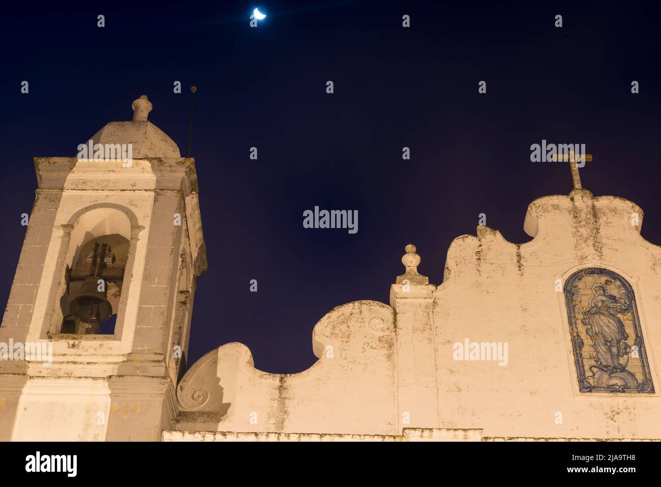 Der Mond über dem beleuchteten oberen Teil der Parochialkirche von Nossa Senhora da Lagoa bei Nacht. Monsaraz, Portugal. Stockfoto