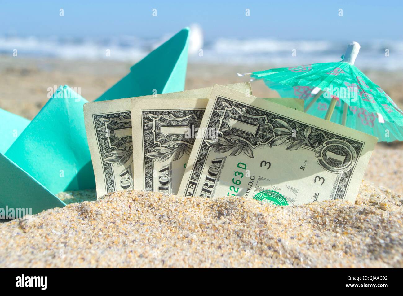 Grünes, kleines Papierboot, eine halbe Papierrechnung für drei Dollar und ein Papiercocktail-Regenschirm am Sandstrand am sonnigen Sommertag. Konzept der Reise Träume Ausgaben Geld finanzieren Tourismus Erholung Urlaub Stockfoto