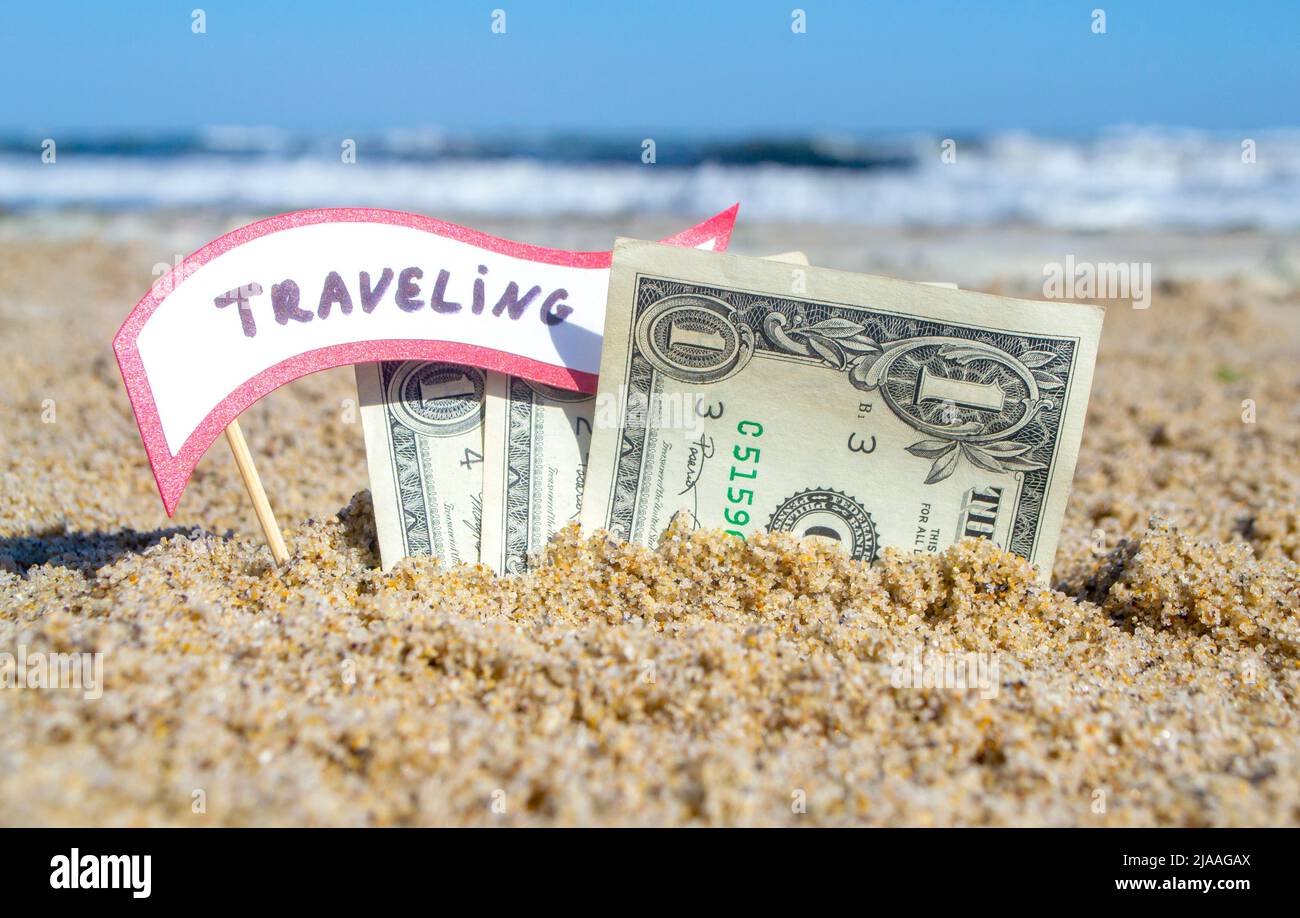 Papierrechnungen ein Dollar und kleiner Stock mit Papier Sprechblase mit Wort Reisen in Sand Strand Hintergrund Meer Nahaufnahme sonnigen Sommertag begraben. Konzept Geld Reise Tourismus Urlaub Urlaub entspannen. Stockfoto