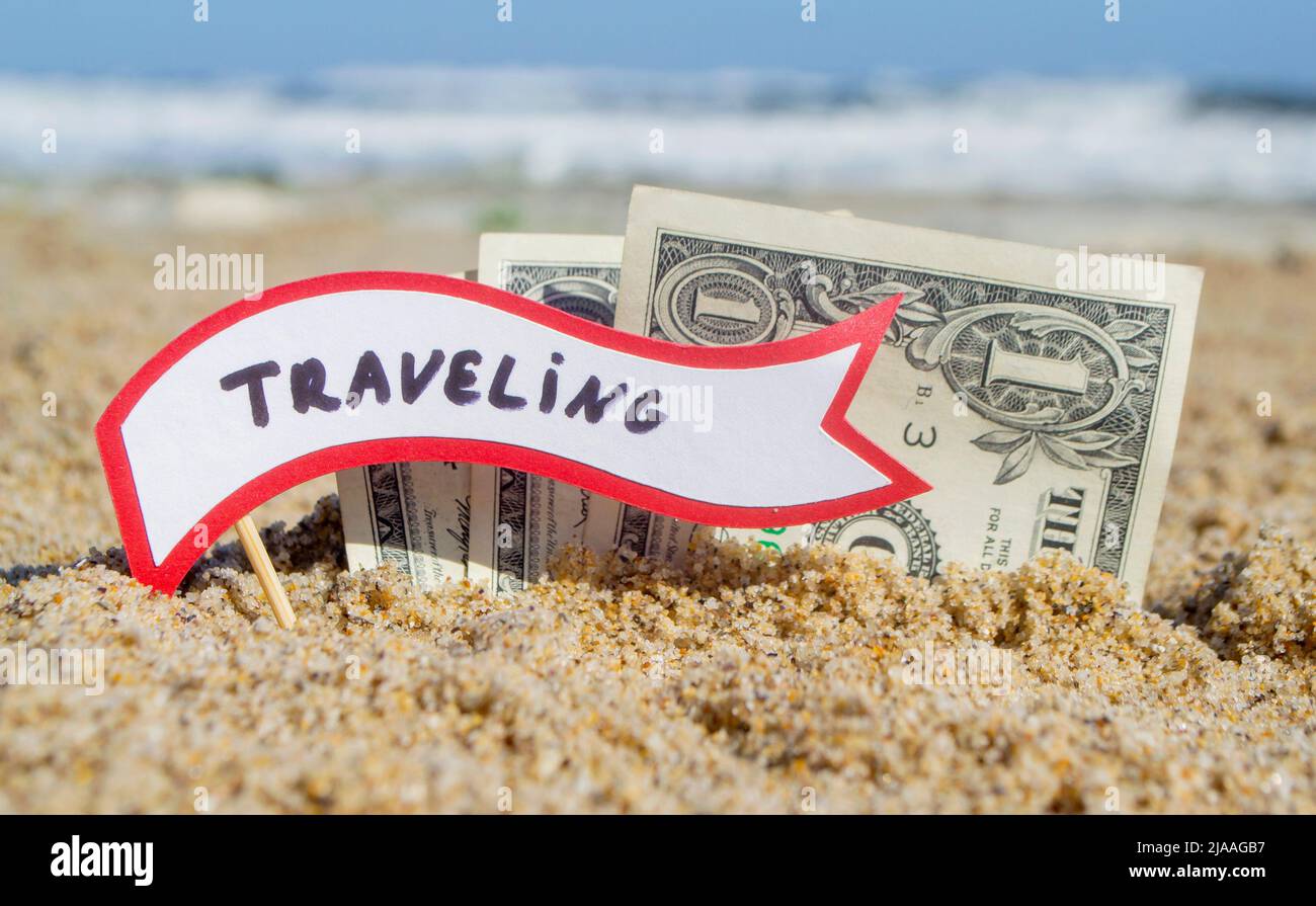 Papierrechnungen ein Dollar und kleiner Stock mit Papier Sprechblase mit Wort Reisen in Sand Strand Hintergrund Meer Nahaufnahme sonnigen Sommertag begraben. Konzept Geld Reise Tourismus Urlaub Urlaub entspannen. Stockfoto