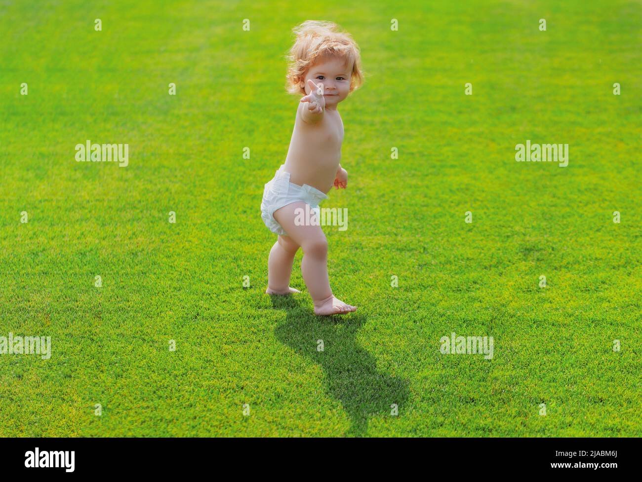 Baby Walking barfuß auf grünem Gras in sonnigen Sommerabend. Kleiner Junge, der im Sommerpark in Windelhosen spielt. Stockfoto