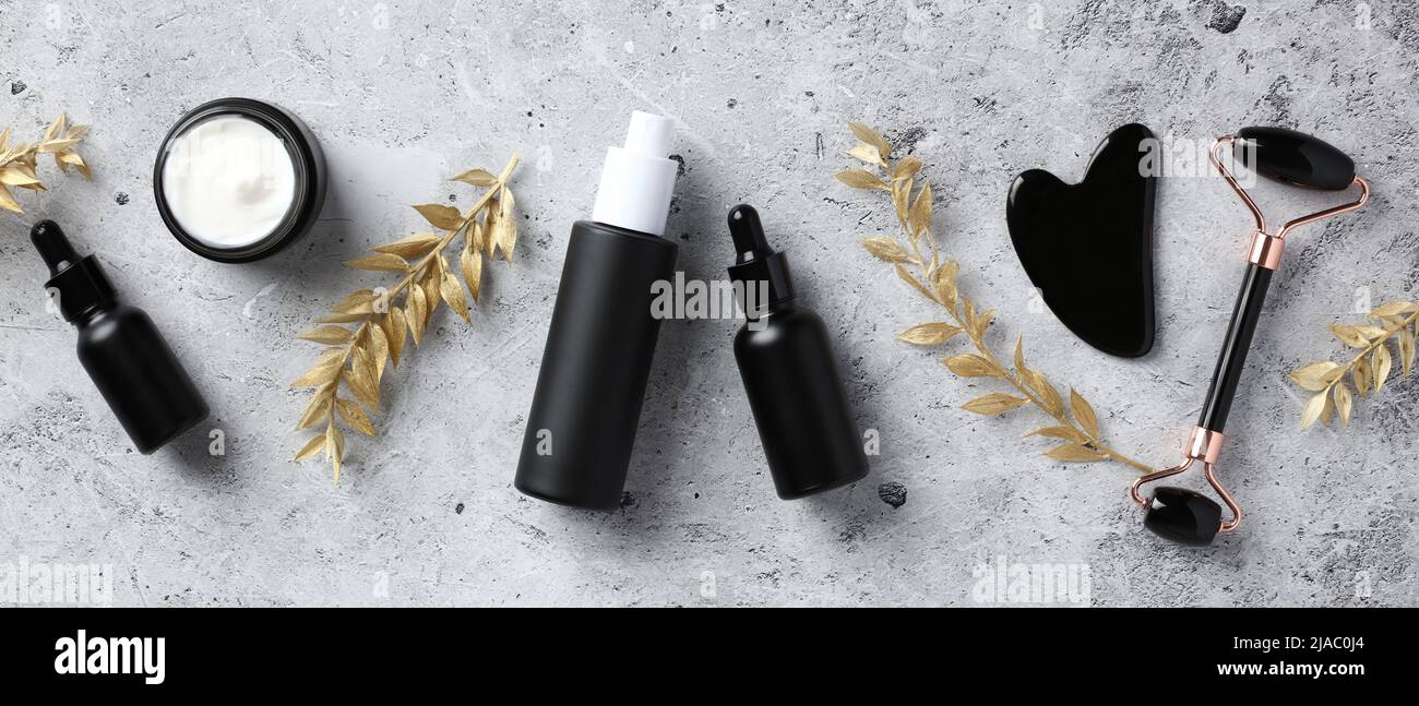 Flache schwarze Obsidian Gesichtswalze, Gua Sha, Serum, ätherische Öle und goldene Zweige auf Steintisch. Luxus-Kosmetik-Set für die Hautpflege. Stockfoto