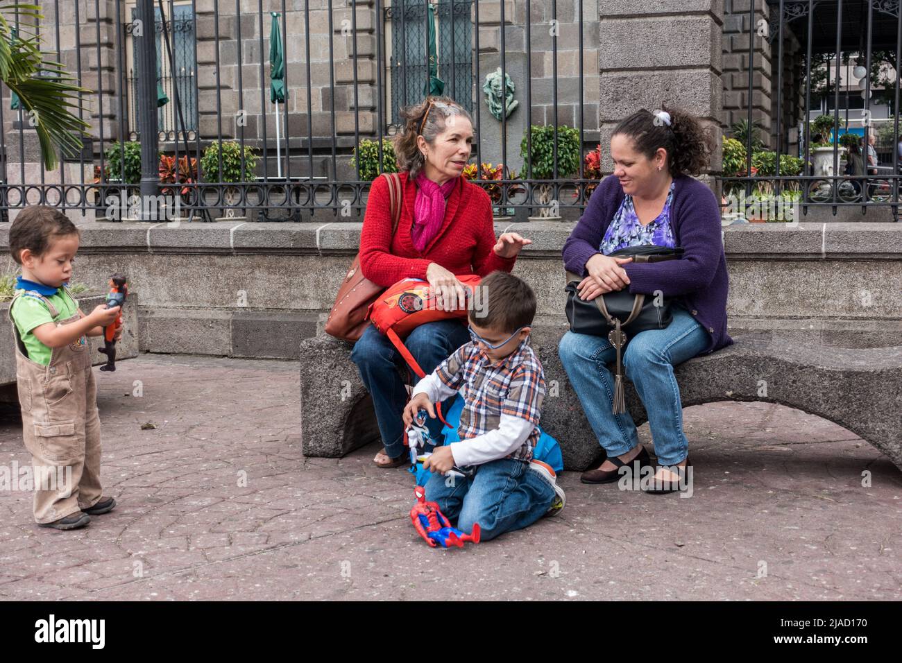 Zwei Frauen unterhalten sich auf einer Parkbank, während zwei Kinder in San José, Costa Rica, mit ihren Spielsachen spielen. Stockfoto