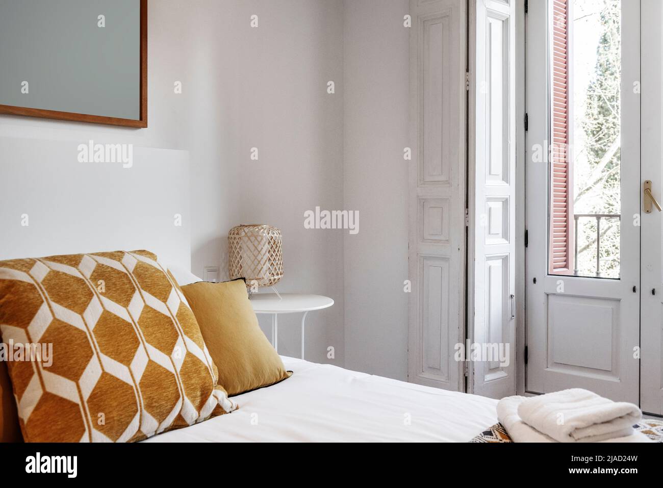 Kopfteil in einem Schlafzimmer mit orangefarbenen Kissen, sauberen weißen Handtüchern und einem Balkon mit Fenstern und weißen Fensterläden aus Holz Stockfoto