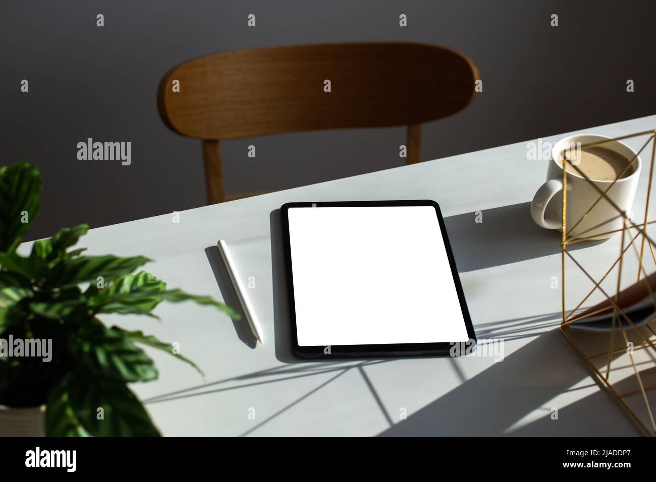 Digitales Tablet mit leerem Bildschirm, Mockup, Stift, Tasse Kaffee, Pflanze auf dem Schreibtisch des Heimbüros. Ästhetischer femininer Arbeitsplatz in Sonnenlichtern Stockfoto