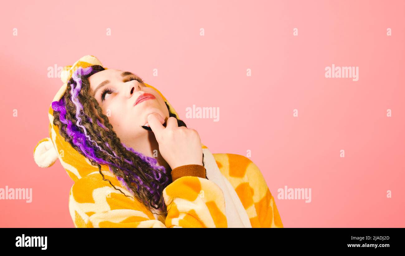 Junge, nachdenkliche Frau in Kigurumi kratzt sich am Kinn und blickt auf rosa Hintergrund. Niedliche lebendige Frau im Schlafanzug mit reflektierenden Gedanken Stockfoto