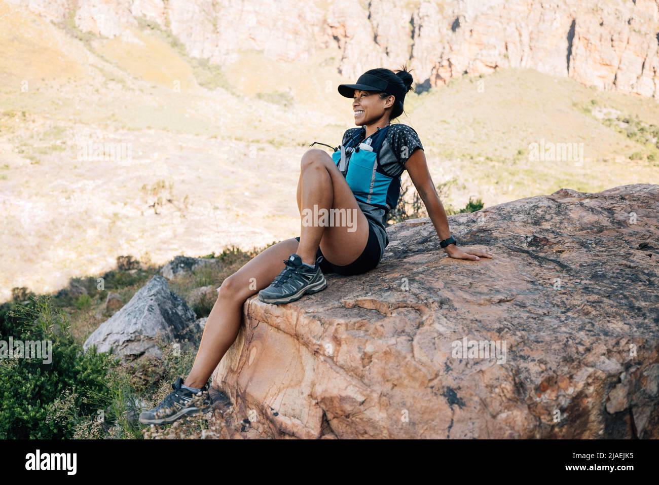 Sportlerin sitzt auf einem großen Stein und nimmt während einer Bergwanderung eine Pause ein. Weibchen genießen die Aussicht im Tal. Stockfoto