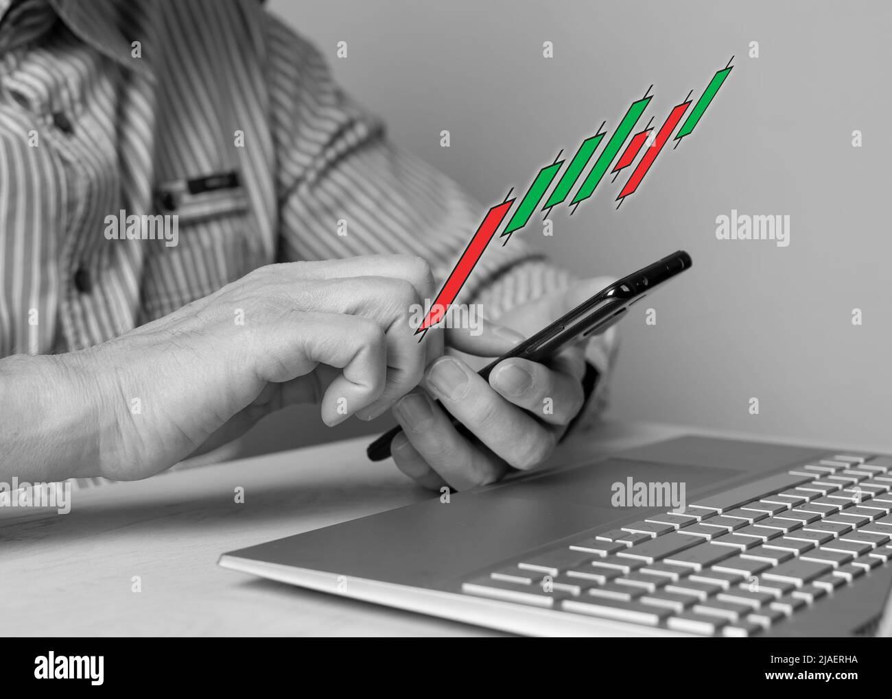Mann mit Telefon in den Händen, der Candlestick-Graph verwendet, um die Kursbewegung zu verfolgen. Börsenanalyse, Investment, Handel, Finanzierungskonzept. Mann sitzt am Tisch mit Laptop. Schwarzweiß. Foto Stockfoto