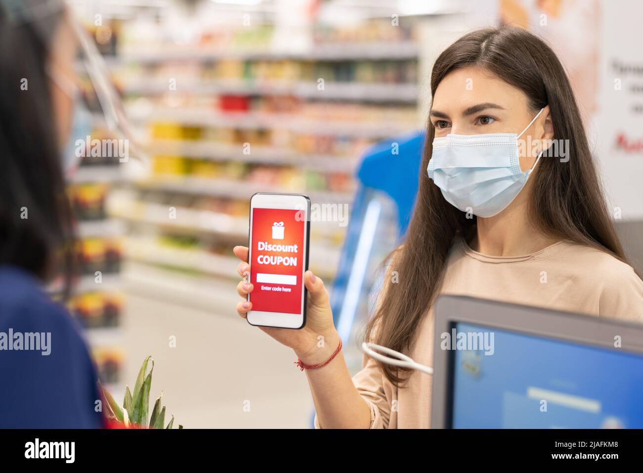 Moderne Frau trägt Maske auf Gesicht mit digitalen Rabatt-Gutschein Auf ihrem Smartphone im Supermarkt Stockfoto