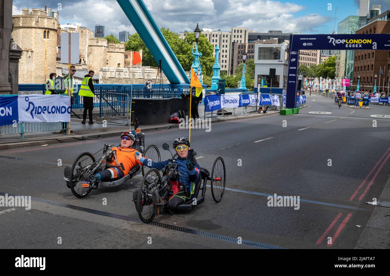 Zwei männliche behinderte Radfahrer, die auf rekumbanten Fahrrädern fahren und am RideLondon 2022 teilnehmen, halten sich nach dem Überqueren der Ziellinie auf der Tower Bridge in die Hände Stockfoto