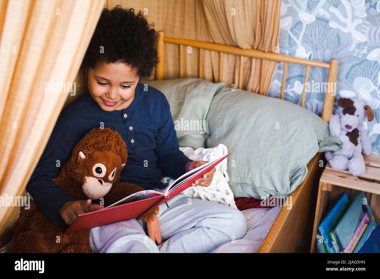 Junge liest das Buch, während er zu Hause mit Spielzeug auf dem Bett sitzt Stockfoto