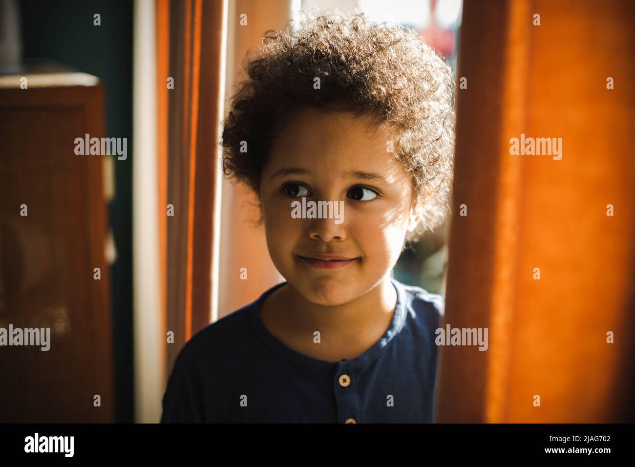 Beschaulicher Junge mit lockigen Haaren, der zu Hause hinter einem Vorhang steht Stockfoto