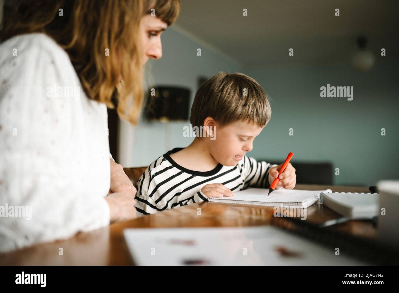 Junge mit Down-Syndrom, der im Buch schreibt, während er von der Mutter am Tisch sitzt Stockfoto