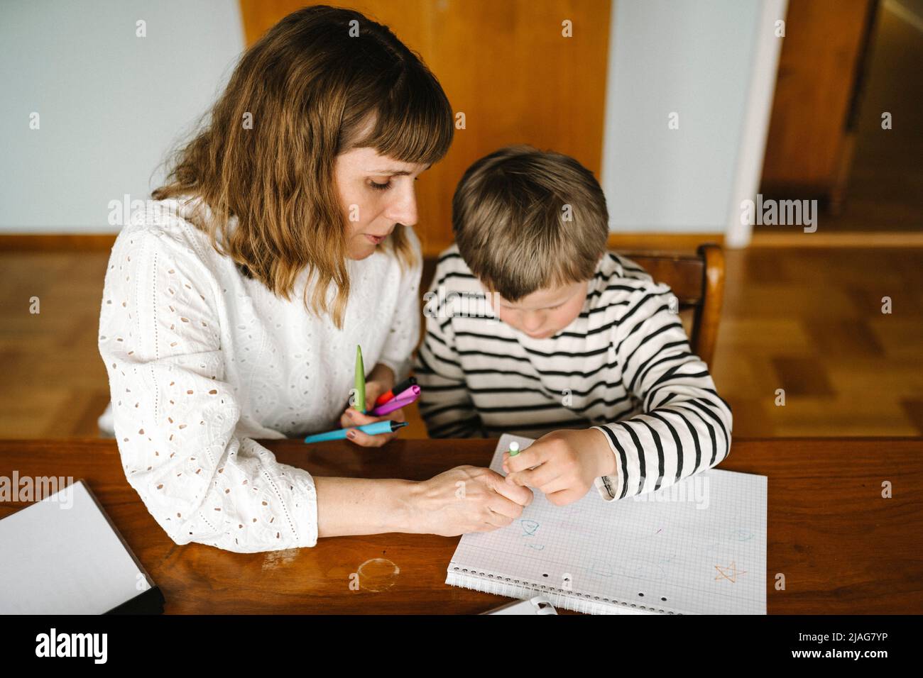 Aus einem hohen Winkel betrachtet, wie die Mutter dem Sohn mit Down-Syndrom beibringt, ein Buch am Tisch zu zeichnen Stockfoto