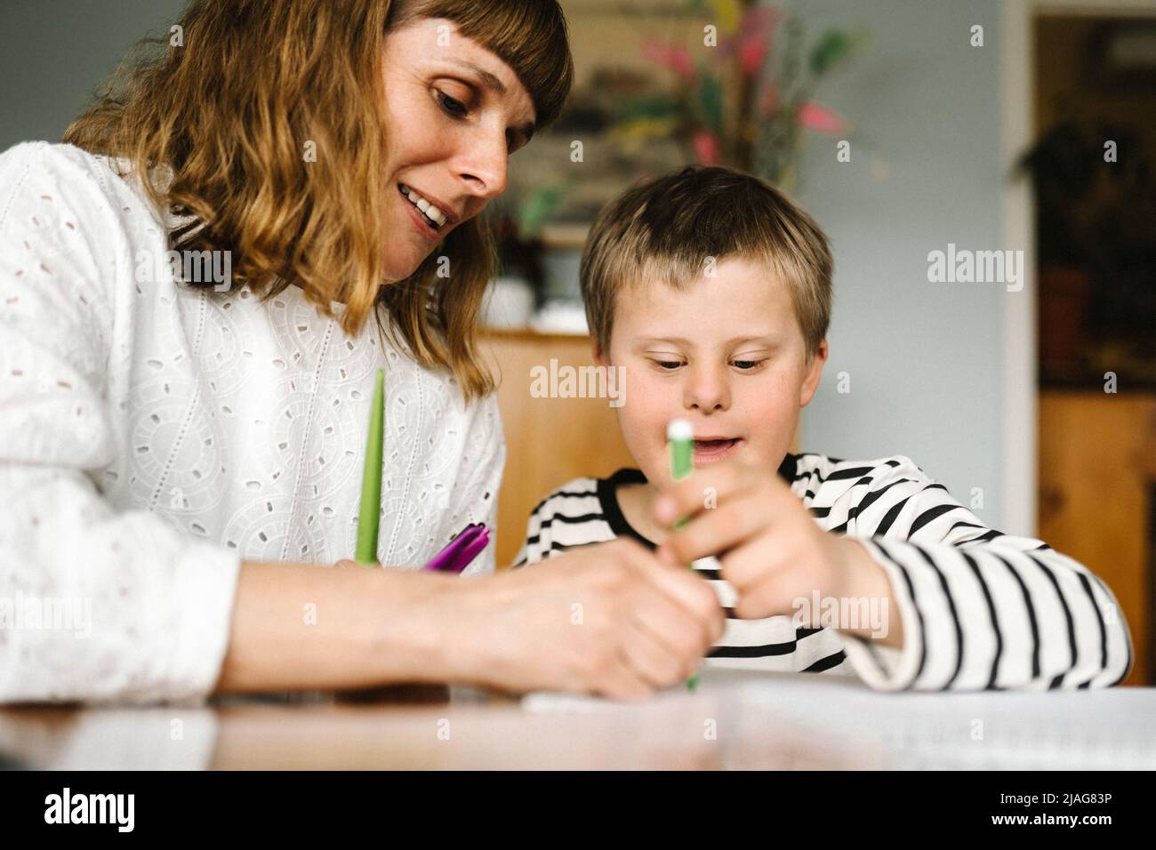 Lächelnde Mutter lehrt Sohn mit Behinderung, zu Hause ein Buch zu zeichnen Stockfoto