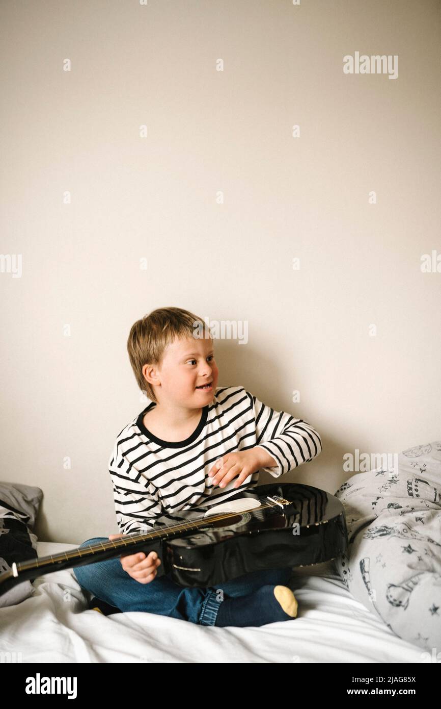 Junge mit Behinderung, der Gitarre trainiert, während er zu Hause auf dem Bett sitzt Stockfoto