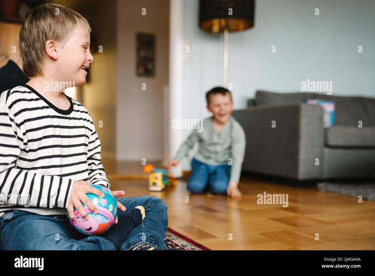 Glücklicher Junge mit Down-Syndrom mit Ball sitzt auf dem Boden im Wohnzimmer Stockfoto