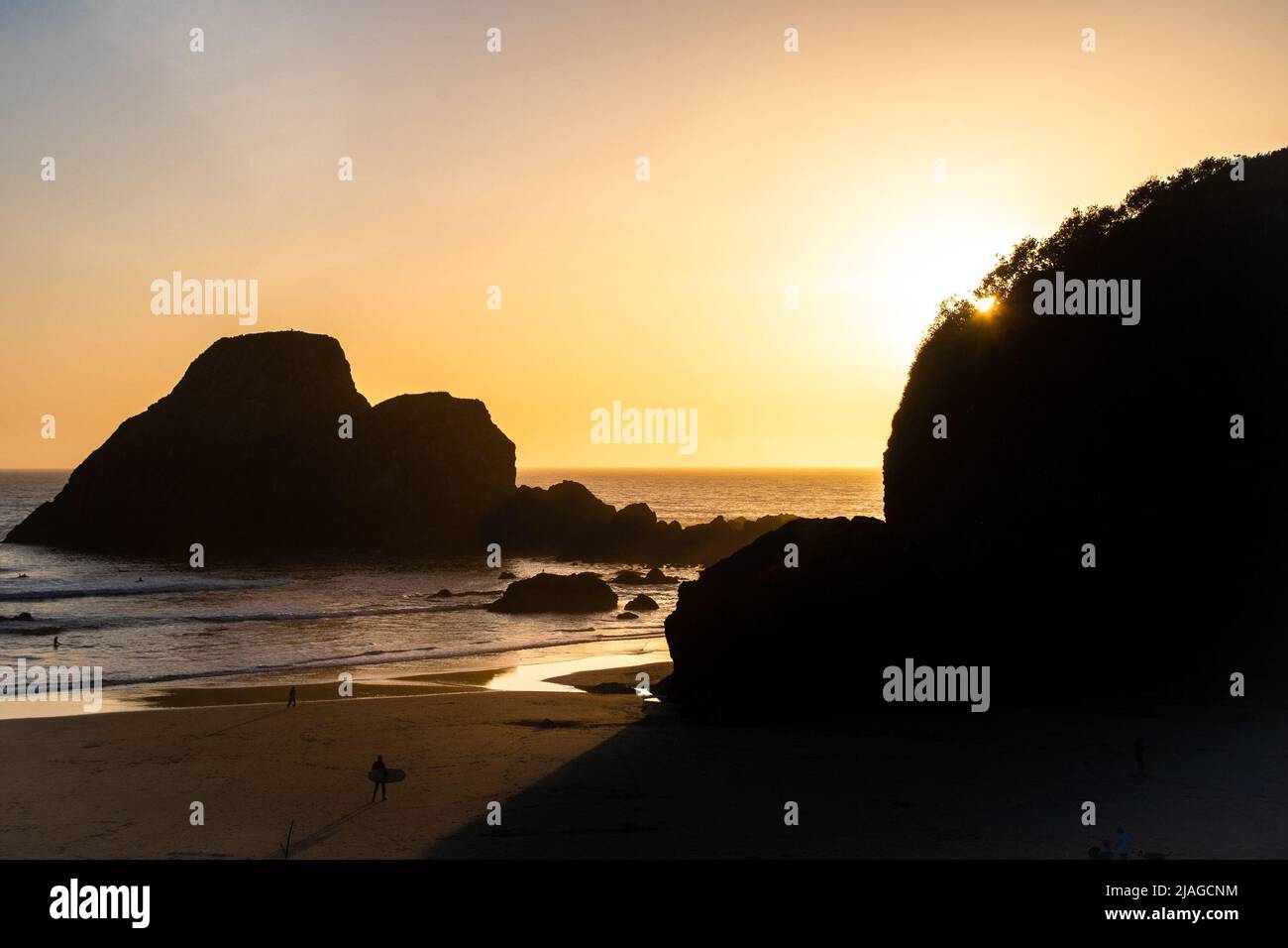 Ein einläufiger Surfer, der in Richtung des Sonnenuntergangs geht, während sich große Schatten ihm vom Sonnenuntergang nähern. Stockfoto