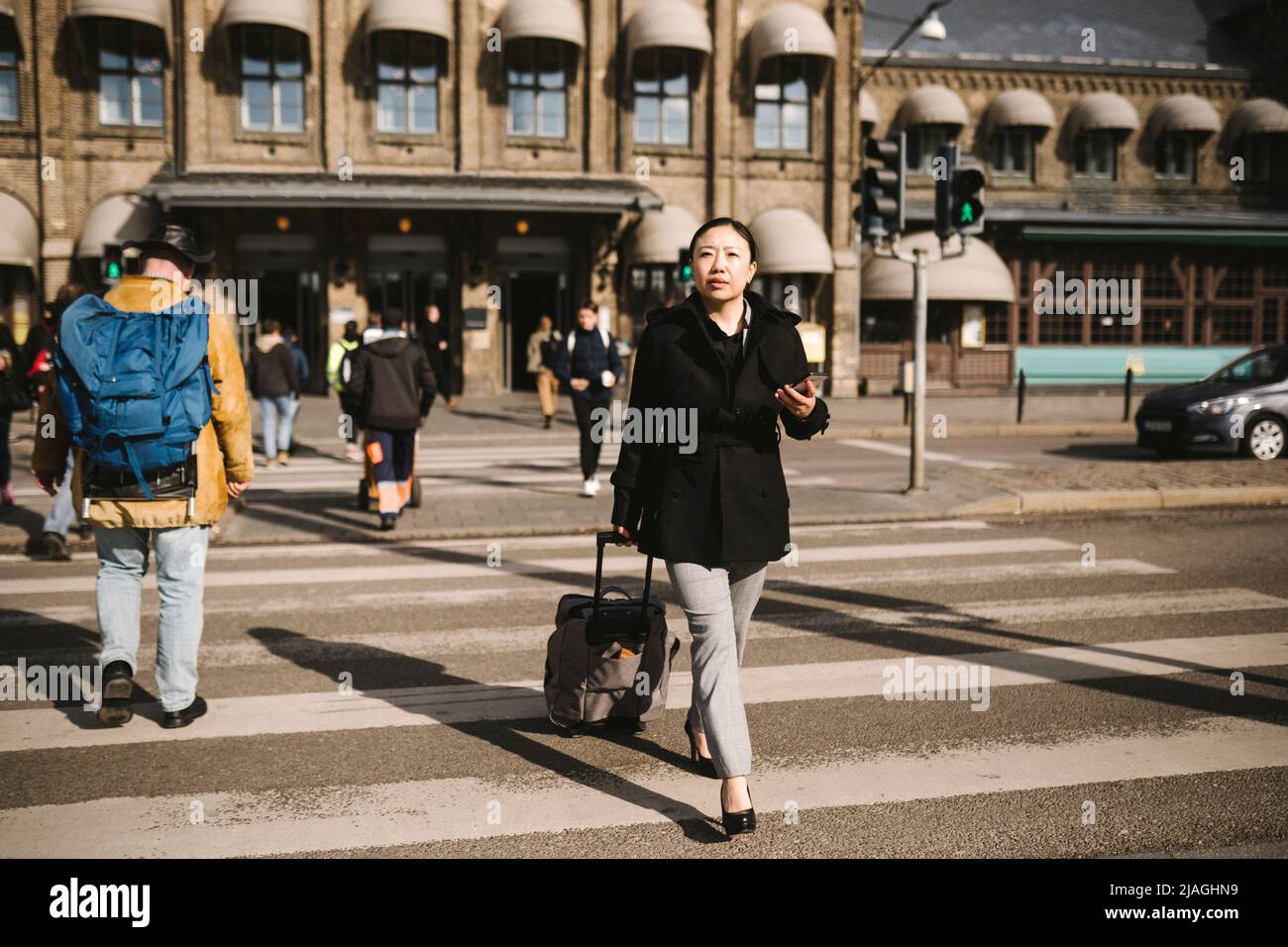Eine Geschäftsfrau zieht ihr Gepäck, während sie an einem sonnigen Tag die Straße in der Stadt überquert Stockfoto