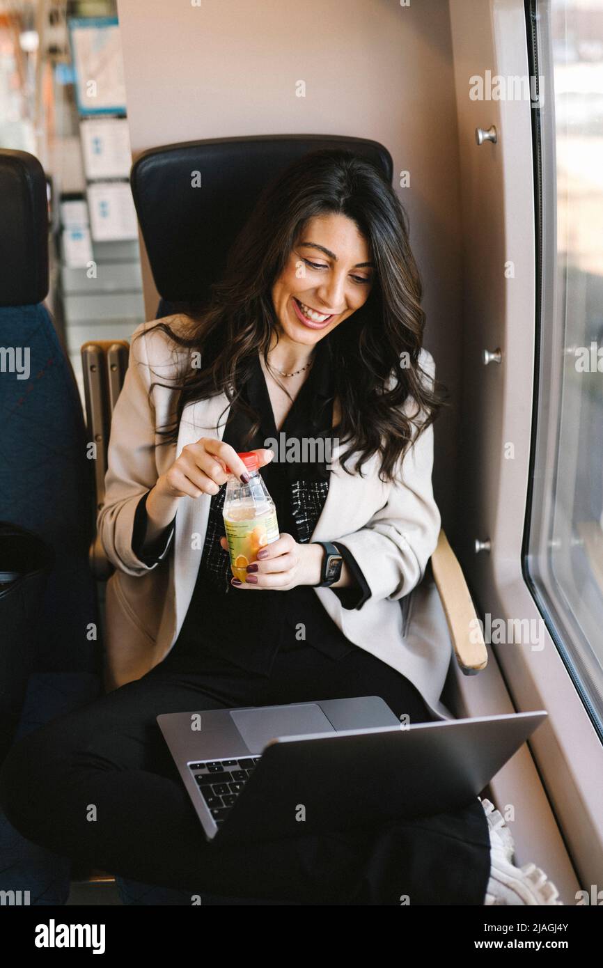 Glückliche Geschäftsfrau, die im Zug eine Trinkflasche öffnete, während sie per Videoanruf über einen Laptop telefonieren konnte Stockfoto