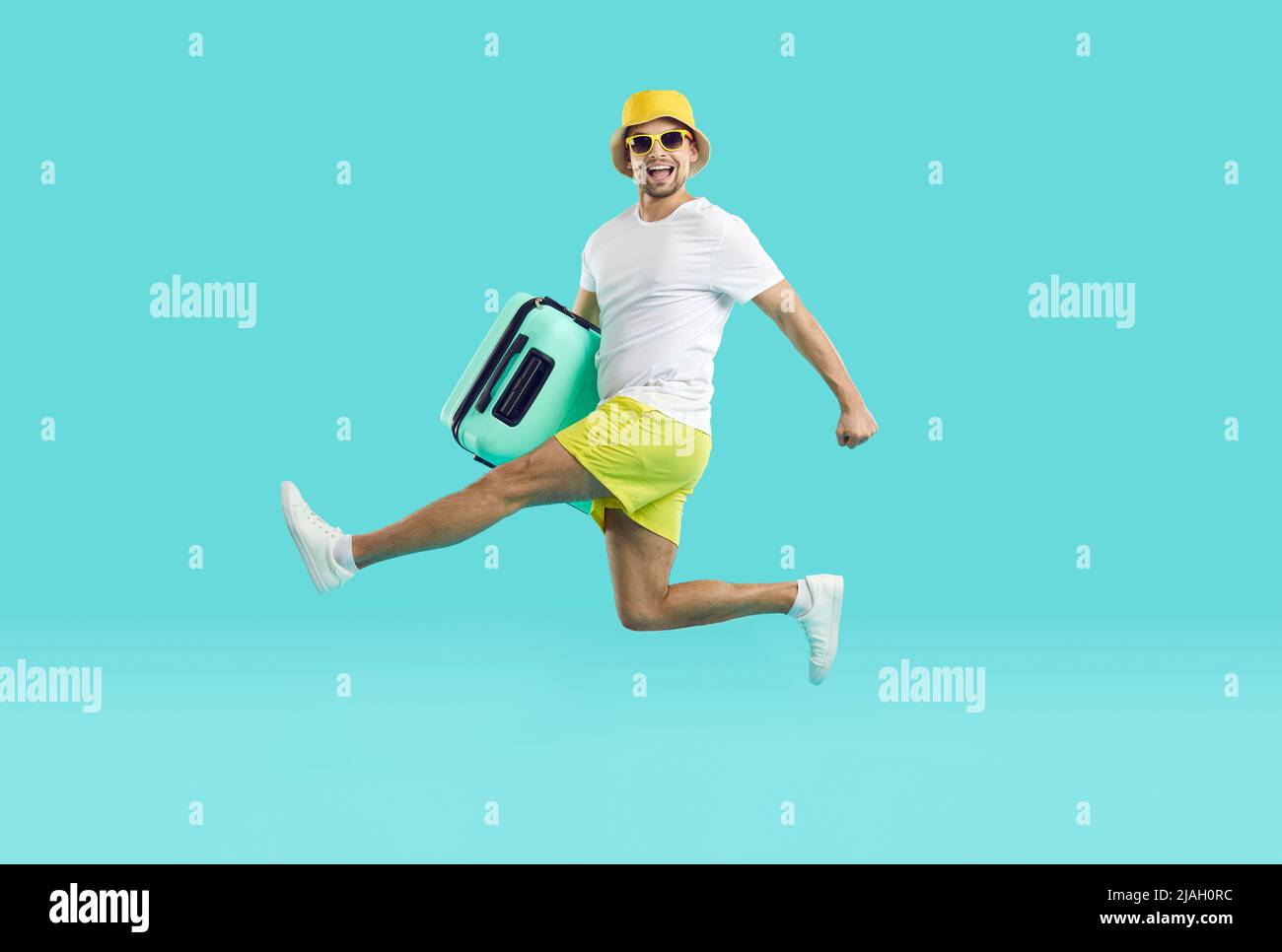 Freudig aufgeregt Kerl in Sommerkleidung läuft und springt mit Koffer auf hellblauem Hintergrund. Stockfoto