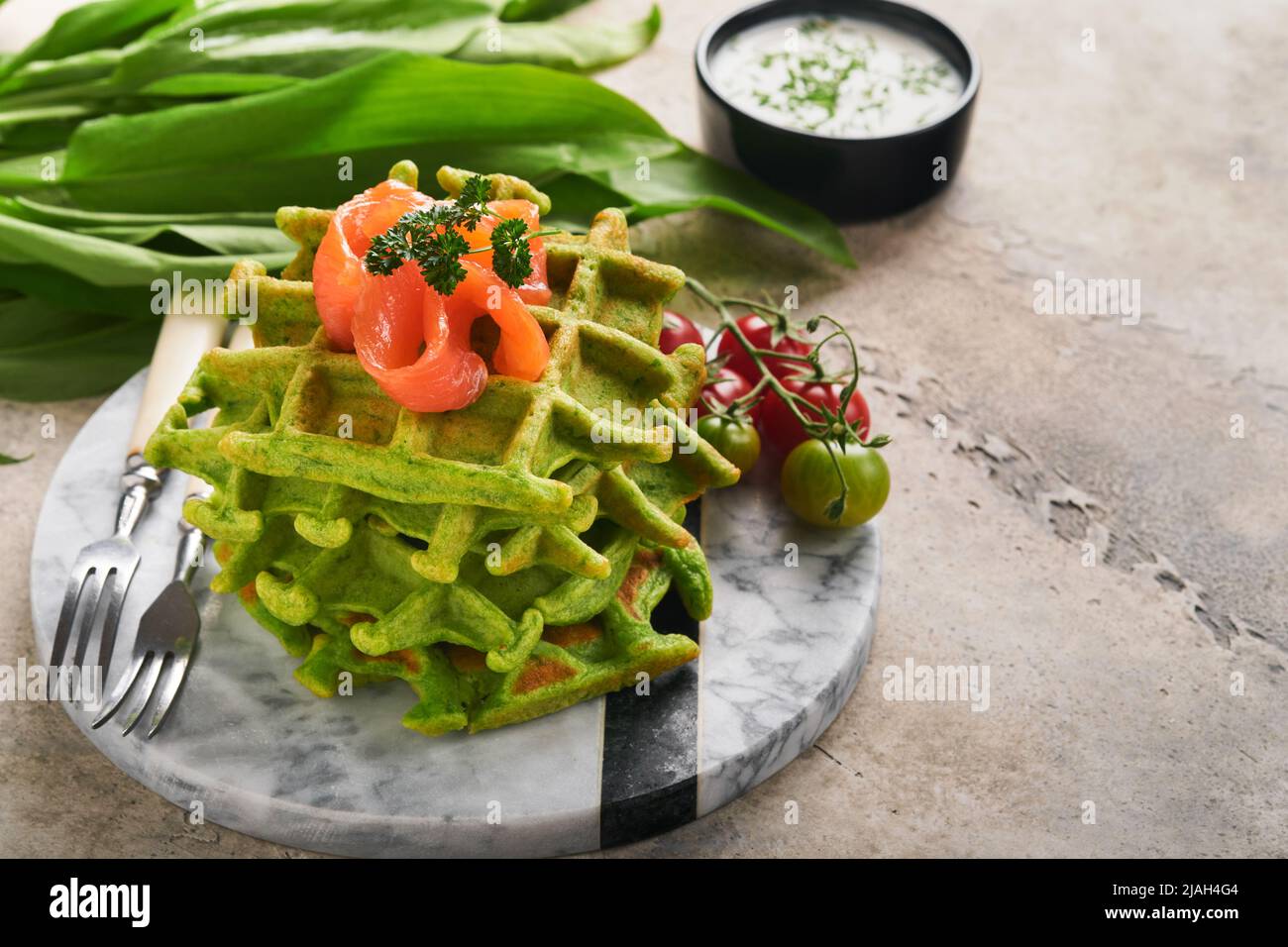 Grüne belgische Waffeln. Spinat- oder Bärlauch- oder Pesto-Waffeln mit rotem Lachs und Sahnesauce auf grauem Beton-Tischhintergrund. Köstliches Frühstück, Stockfoto