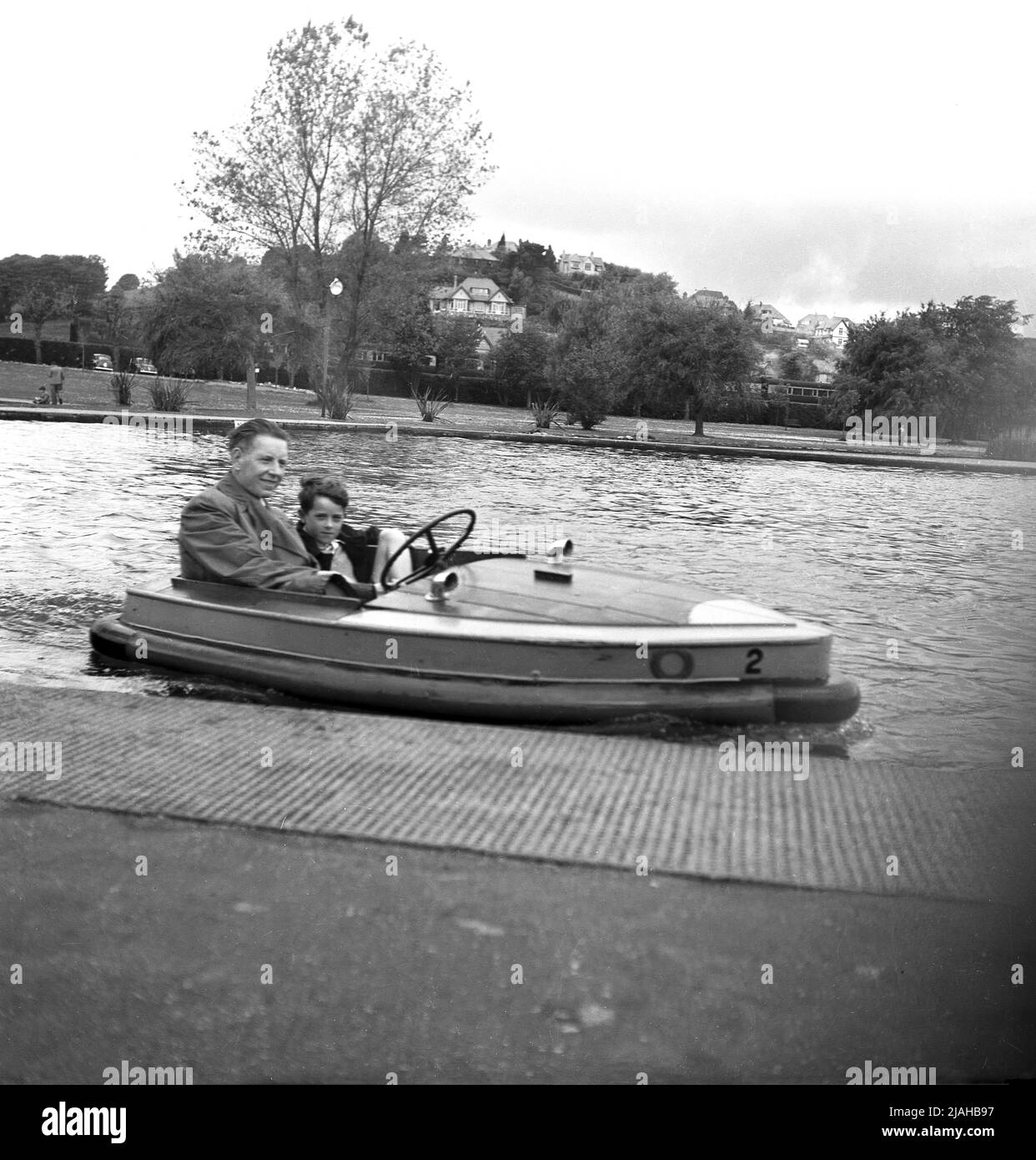 1950s, historisches Großbritannien der Nachkriegszeit, auf einem Bootssee in einem Park, ein Vater mit seinem jungen Sohn zusammen, in einem kleinen hölzernen elektrischen Vergnügungsboot der Zeit. Um die Wende des 20.. Jahrhunderts begannen Bootsseen als eine Form der Freizeit für Menschen, die in Städten leben, gebaut zu werden. Anfang 1930s wurden Ruderboote auf einigen von ihnen durch Schiffe mit neuer Technologie ersetzt, kleine Elektromotorboote mit einer Geschwindigkeit von 4mph, die erste war die "Glida", im Battersea Park im Jahr 1929. Verschiedene Versionen dieses kleinen Elektromotorbootes wurden eingeführt und blieben bis zum 1960s im Einsatz. Stockfoto
