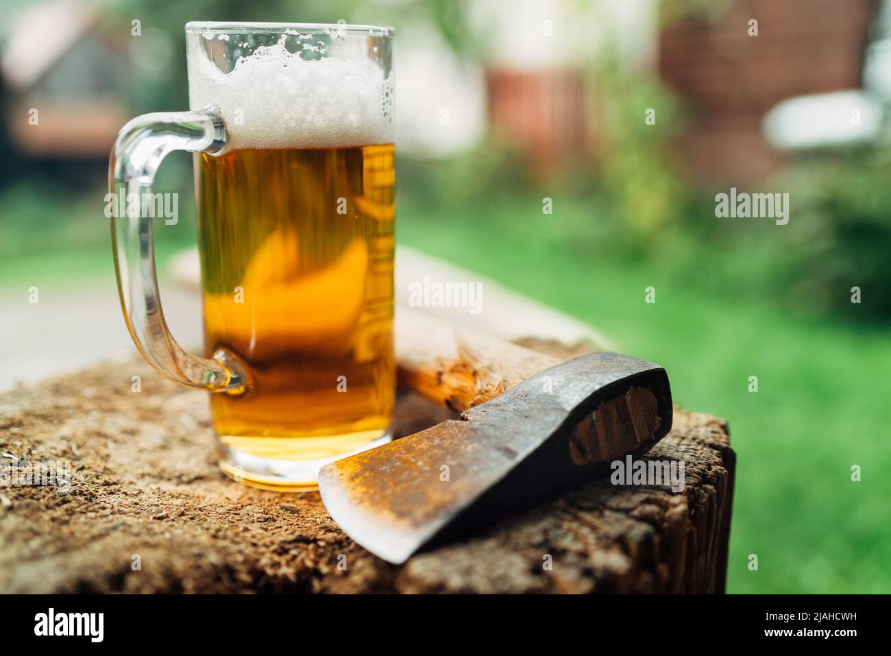 Standbild eines Glases frischen kalten Bieres auf einem Holzstummel neben der Axt. Ländliches Konzept, Männlichkeit, grobes, raues Lifestyle-Konzept in der Wildnis. Stockfoto