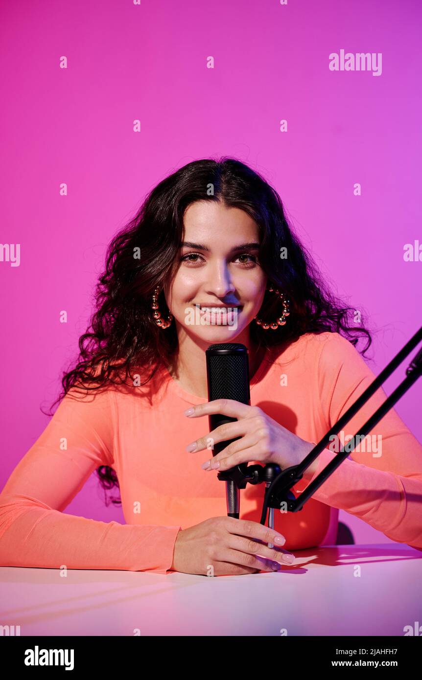 Vertikales Porträt einer fröhlichen Influencerin, die im Studio am Schreibtisch sitzt, in pinkem Neonlicht, das im Mikrofon für ASMR vlog flüstert Stockfoto