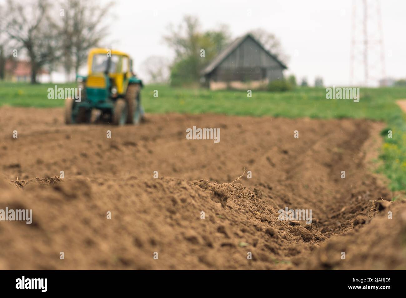 Pflügen und Aussaat des Bodens mit einem alten Traktor auf einem landwirtschaftlichen Feld im Frühjahr, mit alten traditionellen Holzhäusern im Hintergrund, unten Stockfoto