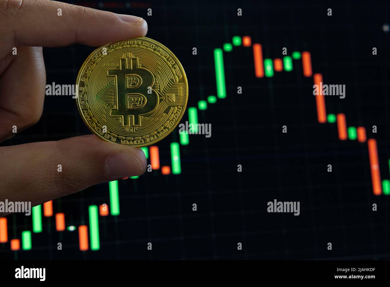 Bitcoin oder Kryptowährung. Foto des Konzepts „Geldverlust auf Kryptowährungsmärkten“. Der Mensch hält einen Bitcoin gegen die Charts. Selektiver Fokus auf den Vordergrund Stockfoto