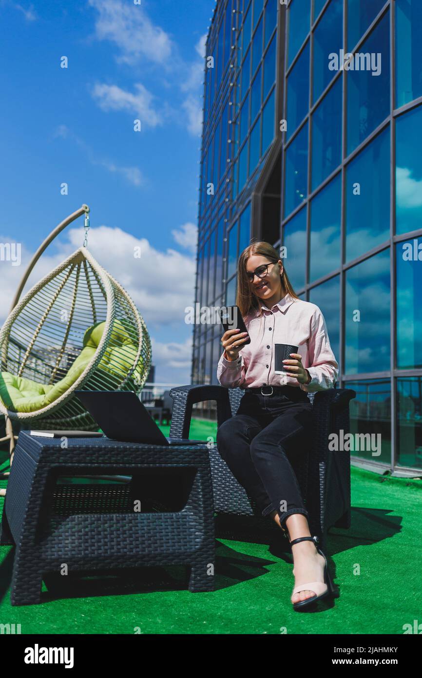 Schöne Frau in klassischer Brille und trendigem Outfit, die eine Nachricht auf einem Mobiltelefon liest und neben einem modernen Glasgebäude steht. Stockfoto